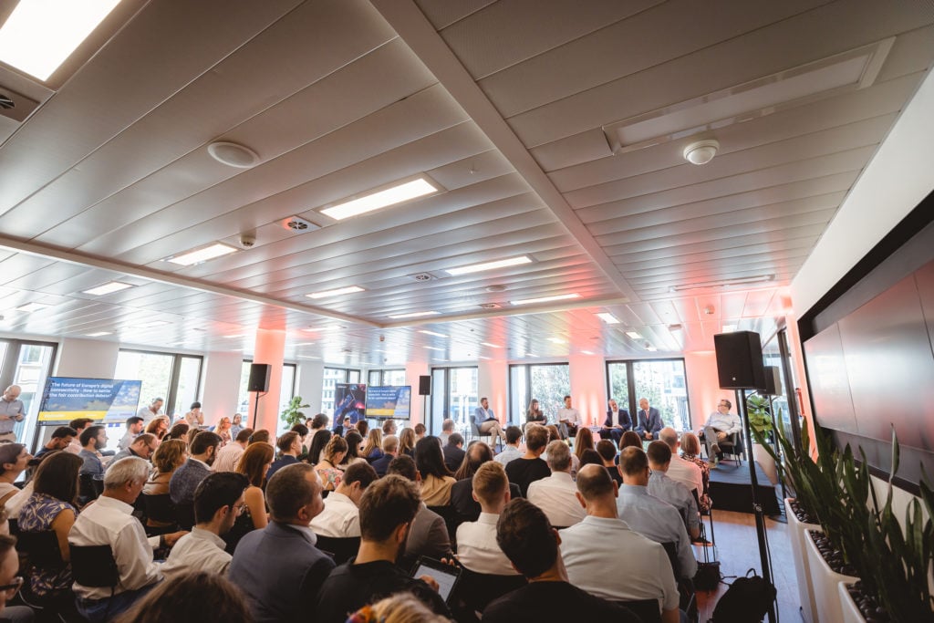 A large group of people sits in rows facing a panel of speakers at the front of a bright, modern conference room. Tall windows let in daylight, and screens display presentation slides. The atmosphere is attentive and professional.