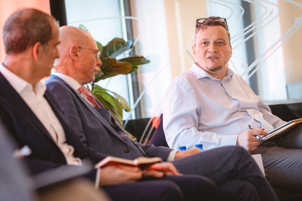 Three men in business attire sit on a panel in a brightly lit room, two holding notebooks while the third, in a light blue shirt, listens and speaks. They wear headset microphones and appear engaged in discussion, with plants and large windows in the background.