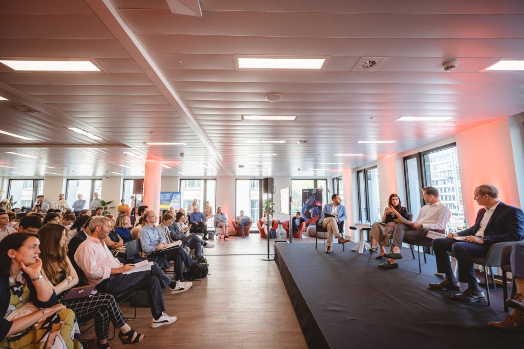 A panel of four people sits on a stage in a modern, well-lit conference room, speaking to an attentive audience. The audience members are seated, some taking notes. Large windows let in natural light, and city buildings are visible outside.