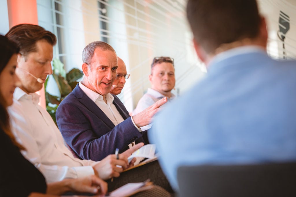 A group of five people, mostly men in business attire, sit in discussion. One man in a dark suit gestures while speaking, holding a notebook. Others listen attentively, one with a pen in hand. The background shows a modern, bright office space.