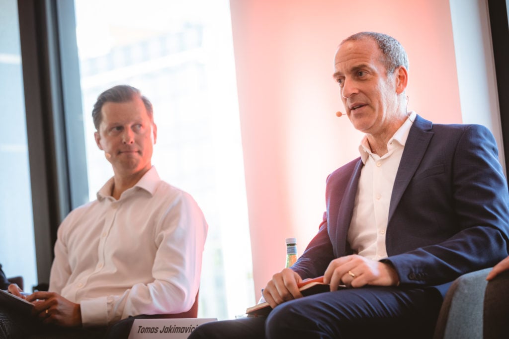 Two men sit side by side in an indoor setting, both wearing business attire and headset microphones. The man on the right gestures while speaking. The background is softly lit with a pinkish hue, and a nameplate reading "Tomas Jokinmovi..." is visible.