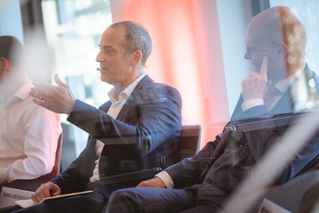 Two men in business attire sit and talk during a panel discussion. One man gestures while speaking, wearing a headset microphone. The second man listens thoughtfully with his hand on his chin. The scene is softly lit with a bright red accent in the background.