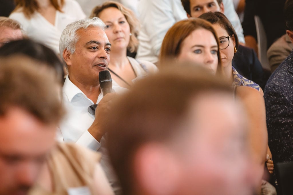 A man with short gray hair, wearing a white shirt, holds a microphone and speaks while seated in a crowd. People around him appear attentive and are dressed in business or casual attire. The scene suggests a conference or discussion event.