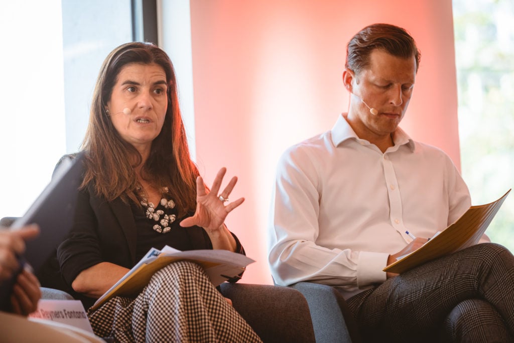 A woman with long brown hair, wearing a black blazer and patterned skirt, speaks passionately with one hand raised. A man next to her in a white shirt and checked pants looks down at his notes. Both wear headset microphones and are seated on stage with papers.