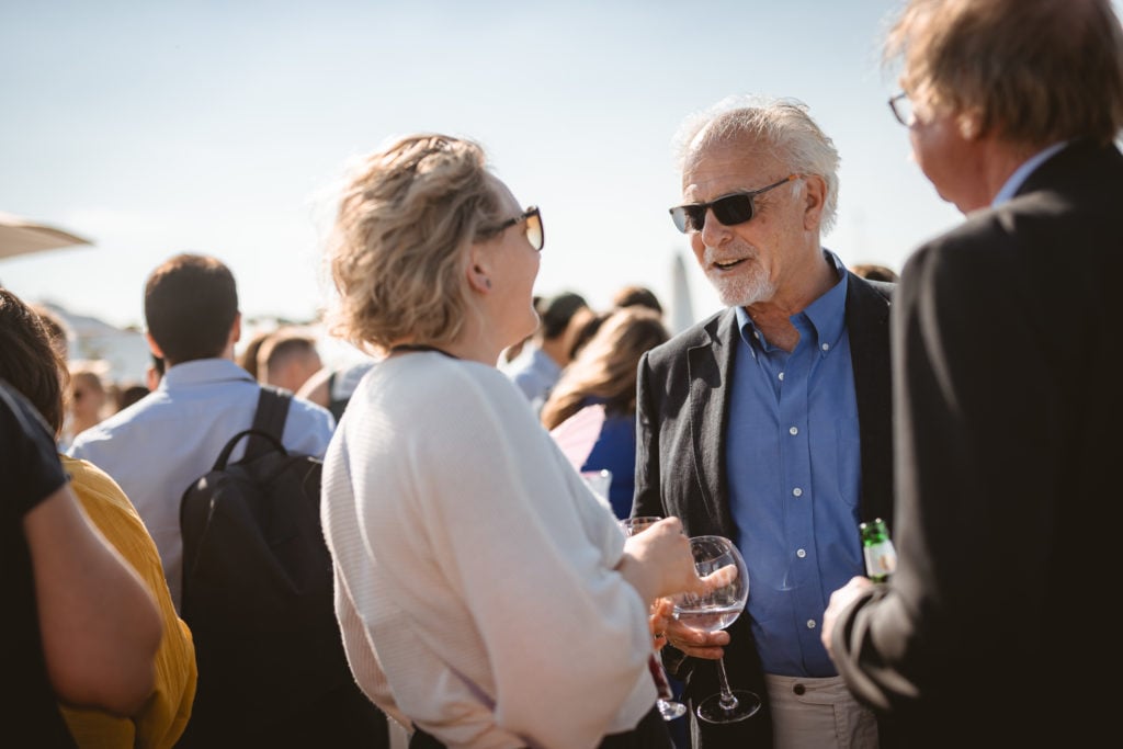 A woman and an older man wearing sunglasses chat and smile at an outdoor social gathering. Both hold drinks. They are surrounded by other people, some blurred in the background, under a bright, sunny sky. The mood appears casual and lively.