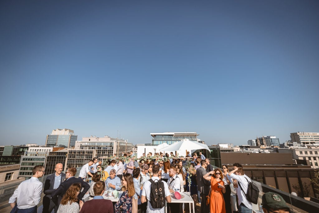 A large group of people in business attire are gathered on a rooftop terrace under a clear blue sky, socializing around tables. Modern city buildings surround the rooftop, and sunlight casts strong shadows across the scene.