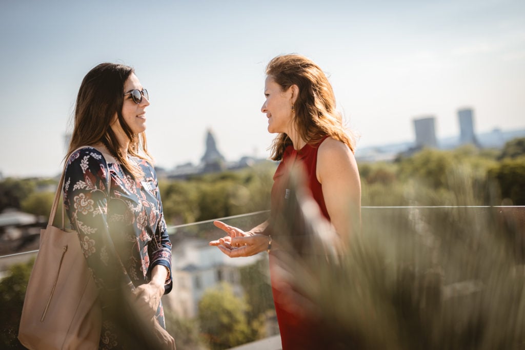 Two women stand outdoors on a sunny day, engaged in conversation. One wears sunglasses and a floral dress, holding a beige bag; the other wears a sleeveless red dress. Trees and buildings are blurred in the background, suggesting a cityscape.