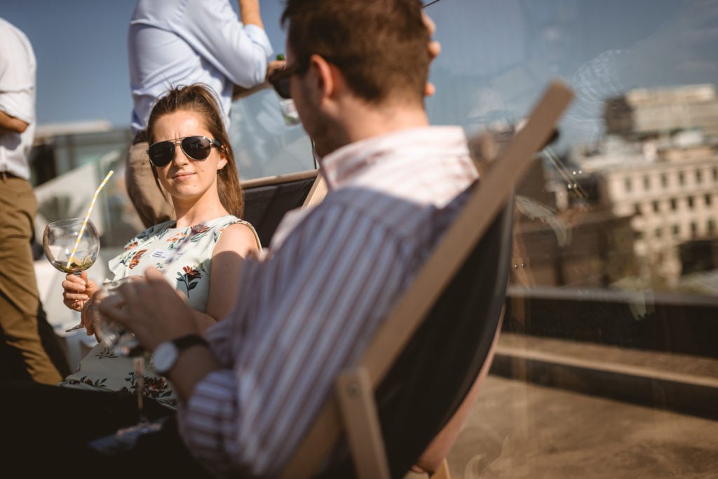 A woman in sunglasses and a floral top sits in a deck chair, holding a drink with a straw, gazing at a man in a striped shirt. They are outdoors on a rooftop with reflections of buildings and people in the glass wall beside them.