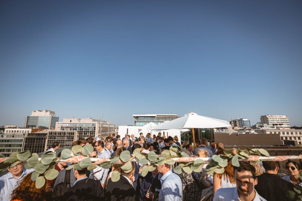 A large group of people attends a rooftop event under a clear blue sky. Some guests are under a white umbrella. City buildings surround the scene. Green leaves strung on a garland hang in the foreground, partially blocking the view.