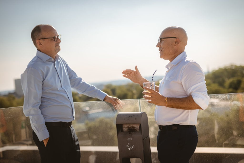 Two middle-aged men wearing glasses and collared shirts talk on a sunny outdoor terrace. One holds a drink and gestures with his hand, while the other stands with hands in his pockets. A small black machine is between them, and trees are visible in the background.