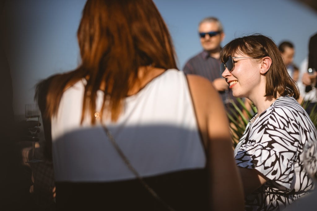 Two women are in focus at an outdoor event. One woman with brown hair and sunglasses smiles, facing right. The other, with long auburn hair, is seen from behind. The background shows people in casual attire, blurred against a clear blue sky.