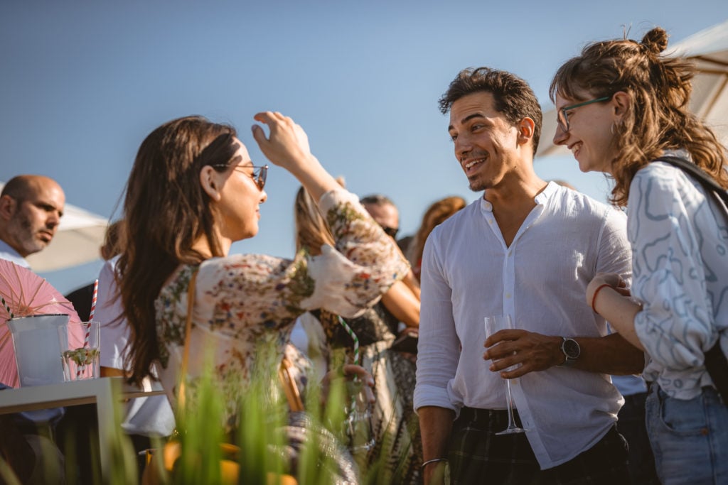Three people stand outdoors, smiling and chatting at a sunny social gathering. The man in the center wears a white shirt and holds a drink, while the two women on either side wear glasses and light tops. Blurred guests and a clear blue sky are in the background.