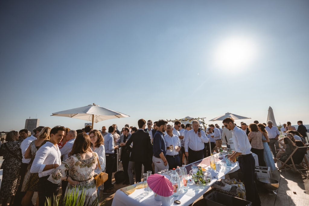A large group of people in semi-formal attire gather around tables with drinks and snacks at a rooftop event on a sunny day. Two large white umbrellas provide shade. The sky is clear and bright, with the sun shining overhead. City buildings are visible in the distance.