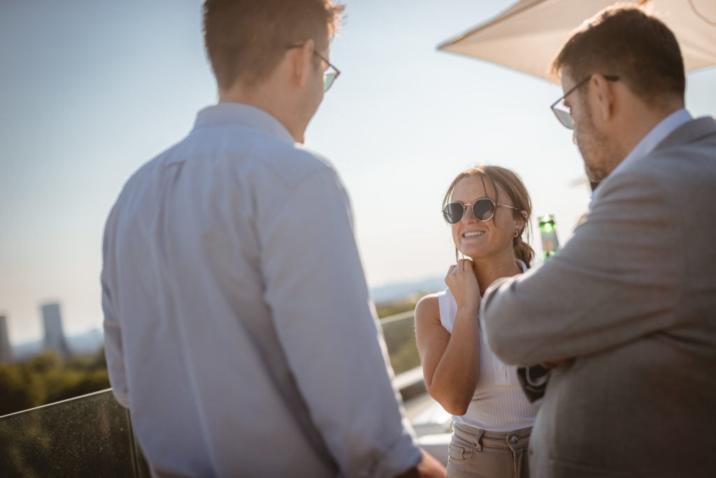 Three people stand outdoors on a sunny day, talking and smiling. One woman in sunglasses faces two men, one holding a drink. They are casually dressed, near a glass railing, with city buildings and trees blurred in the background.