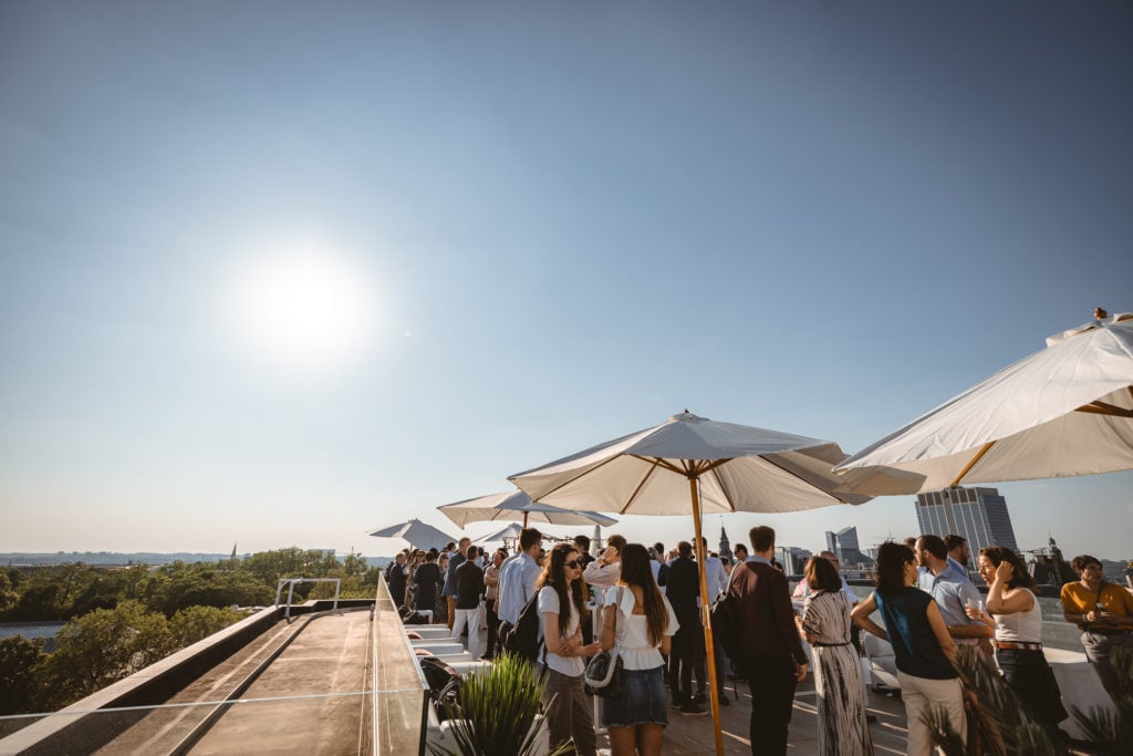 A large group of people socialize on a sunny rooftop terrace with white umbrellas. The crowd is casually dressed, and the scene overlooks trees and city buildings under a clear blue sky. The bright sun shines in the upper left corner.