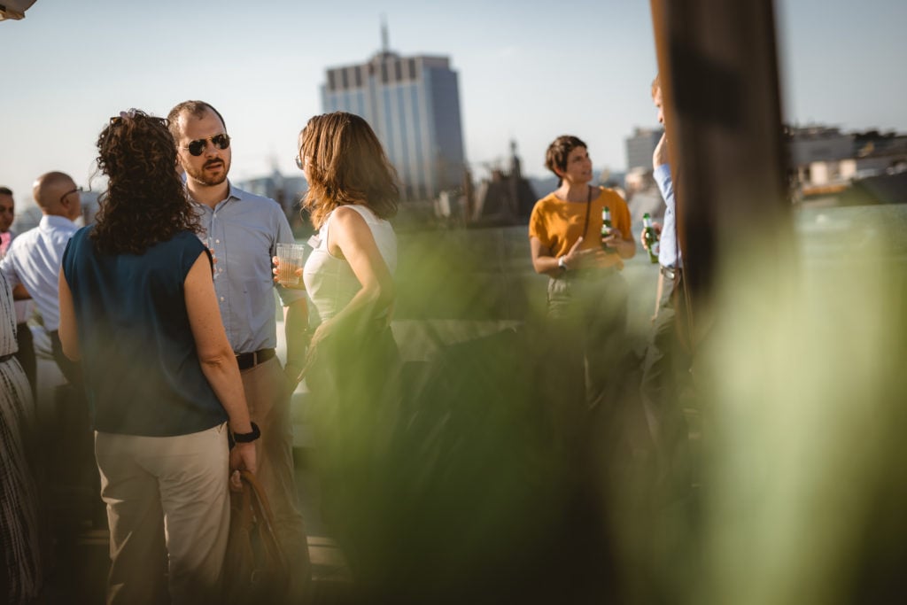 A group of people socialize outdoors on a sunny rooftop with a cityscape in the background. Several individuals stand in conversation, some holding drinks. Green foliage is blurred in the foreground, and a tall building is visible in the distance.