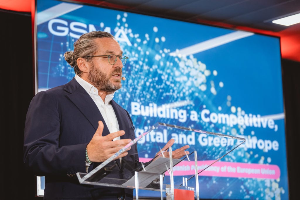 A man with glasses and a beard, dressed in a dark suit and white shirt, speaks at a clear podium. Behind him, a digital screen displays the text "Building a Competitive, Digital and Green Europe" with a blue, tech-themed background.