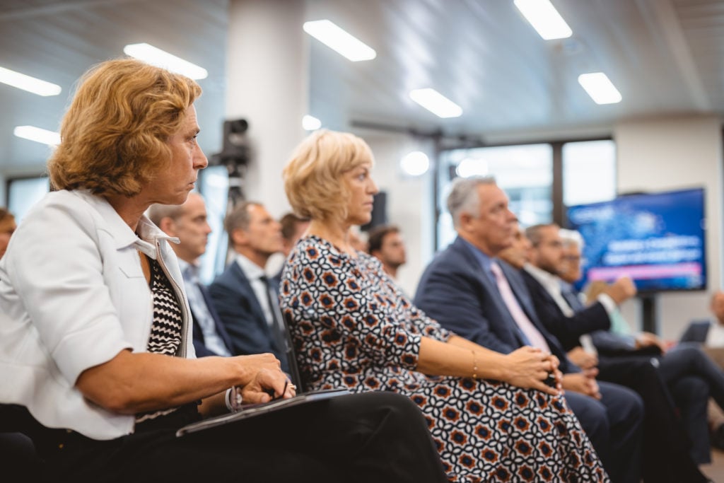A group of professionally dressed adults sits attentively in a conference room. The focus is on two women in the foreground, both listening intently. The background shows other attendees, a large window, and a blurred presentation screen.