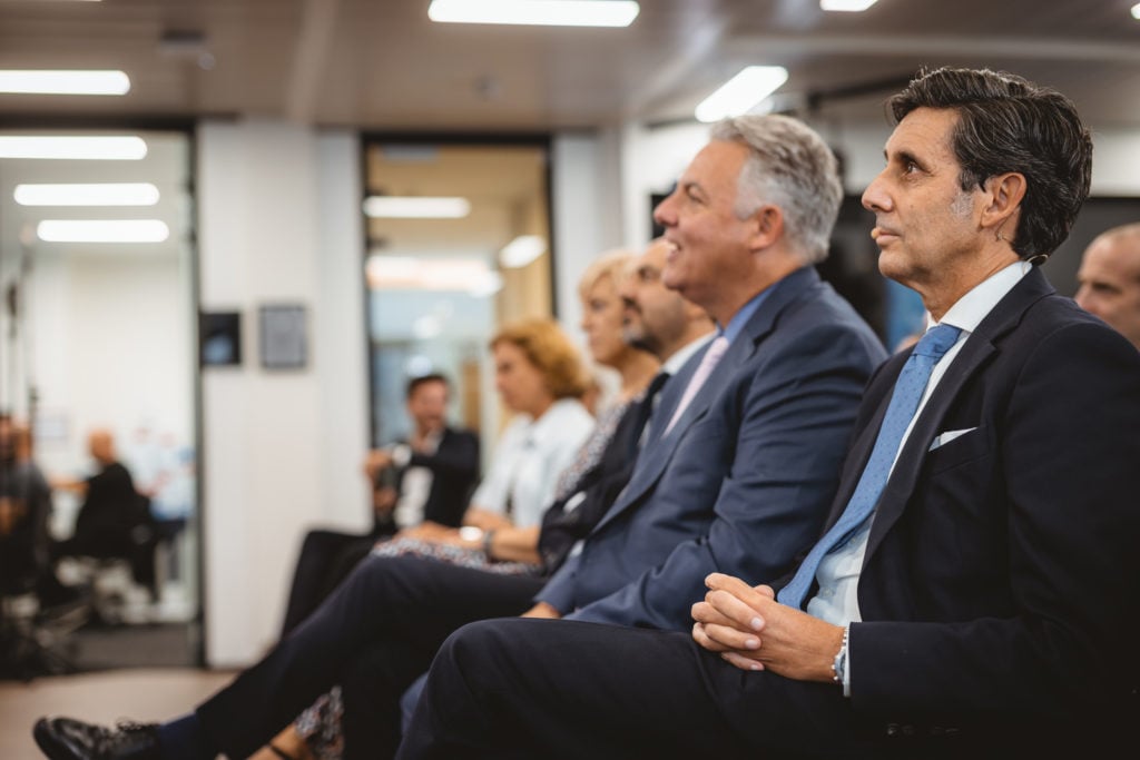 Several formally dressed adults sit attentively in a row at an indoor event or conference. The focus is on a man in a dark suit and blue tie in the foreground, while others, men and women, are slightly blurred in the background. The setting appears professional.