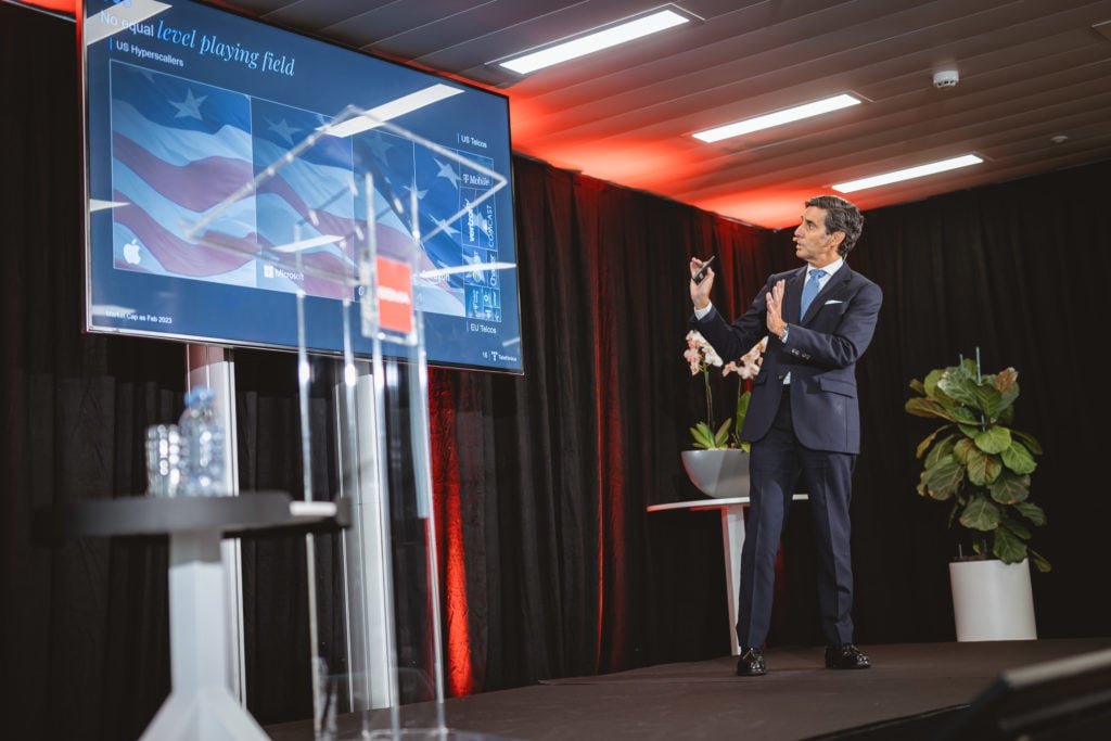 A man in a dark suit gestures while presenting on stage next to a large screen showing graphs and US flag imagery. The stage has bright lighting, a podium, a potted plant, and a water bottle, with a black curtain backdrop and red accents.