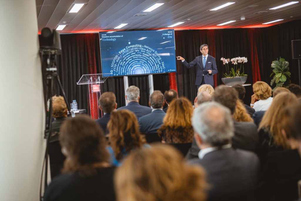 A man in a suit stands on stage, pointing at a large screen displaying a word cloud, addressing an audience seated in rows. The room has black curtains, red lighting, potted plants, and a podium with a microphone.