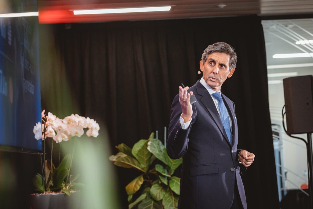 A man in a navy suit and blue tie stands on stage with a headset microphone, gesturing with his right hand. Behind him are large green plants, white orchids, and a black curtain. Soft lighting highlights his focused expression.