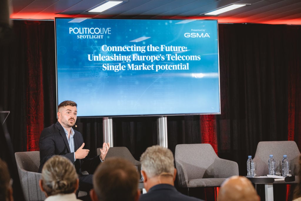 A man in a suit speaks on stage during a panel, gesturing with his hands. Behind him is a large screen displaying the text: “Connecting the Future: Unleashing Europe’s Telecoms Single Market potential.” Three empty chairs and water bottles are visible.
