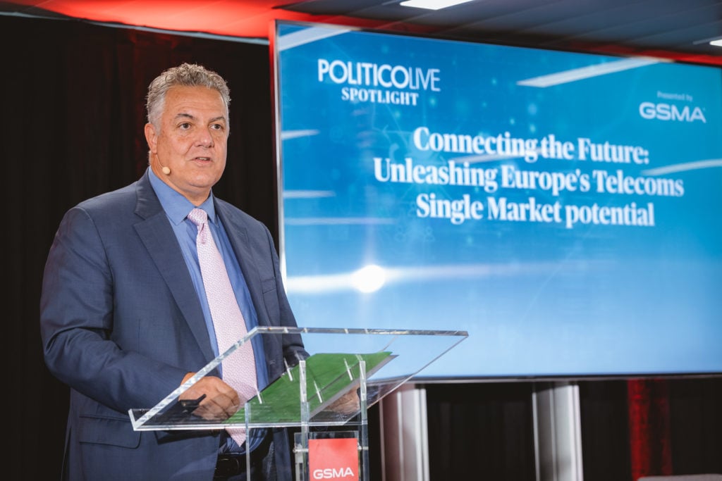 A man in a suit stands at a clear podium with a microphone headset, speaking at an event. Behind him, a large screen displays the text: "POLITICO LIVE SPOTLIGHT: Connecting the Future: Unleashing Europe’s Telecoms Single Market potential.