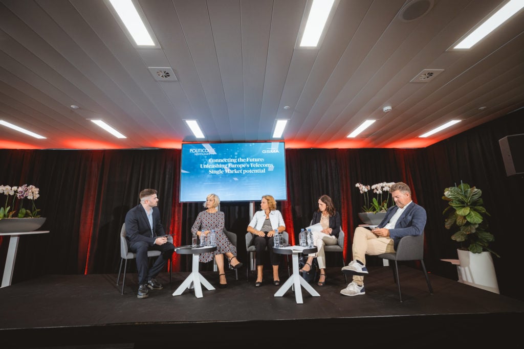Five people sit on stage under warm lighting, engaged in a panel discussion. Behind them, a screen displays the event title. Two women and three men are seated on modern chairs, with small tables and plants around them, and dark curtains as a backdrop.