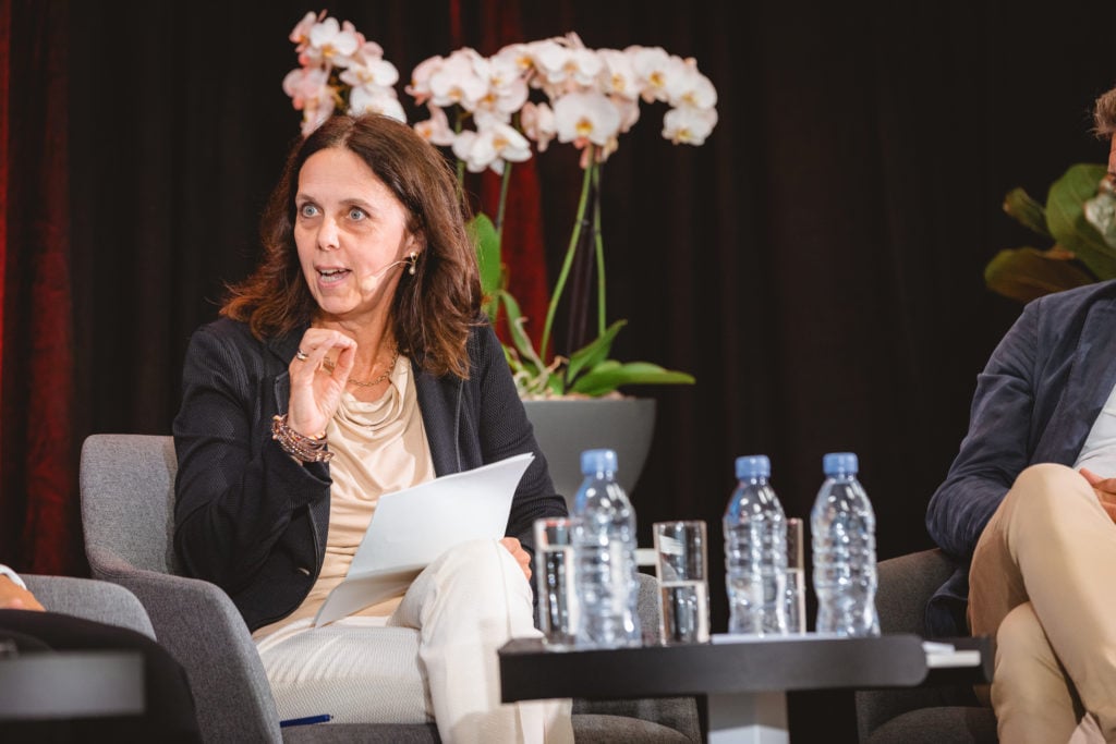 A woman with long brown hair speaks while holding papers onstage in a panel discussion. She wears a beige outfit and black blazer, with a headset microphone. Three water bottles and glasses are on the table. Orchids and dark curtains are in the background.