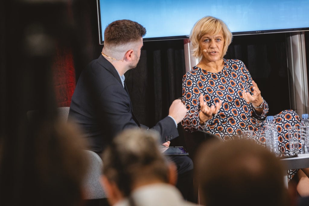 A woman in a patterned dress speaks expressively in a discussion with a man in a dark suit. They are sitting on stage with a screen behind them, engaging with an unseen audience. The foreground is blurred, showing the backs of attendees’ heads.