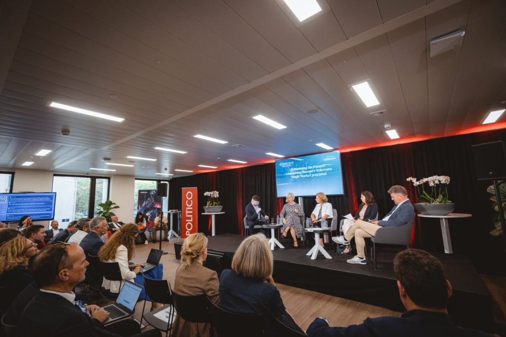 A panel of five people sits on stage in front of an audience at a Politico event. Behind them, screens display discussion topics. The audience is seated, some taking notes or photos. The room is modern, with ceiling lights and large windows on the left.