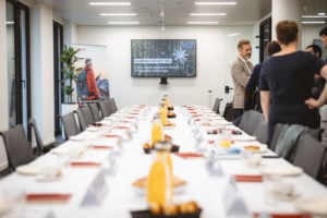 A conference room with a long table set for a meeting, lined with chairs, coffee cups, plates, and pastries. People stand and talk at one end. A large screen displays the words "ISGMA.CONNECTED" and a mountain scene. The room is bright and modern.