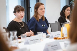 Three women sit at a conference table with name cards, glasses, pastries, and a jug of orange juice. The woman on the left speaks, wearing a black top and gold necklace. Another woman in blue listens attentively; a third woman is further back on the right.