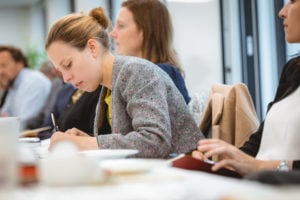 A woman in a gray blazer sits at a table, leaning forward and writing intently in a notebook. Other people, slightly out of focus, sit beside her in a bright conference room, suggesting a group meeting or workshop setting.