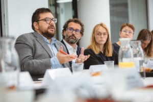 A man in a gray suit speaks and gestures with his hands at a conference table, surrounded by attentive colleagues. Documents, glass pitchers, and cups are visible on the table in a bright, modern meeting room.