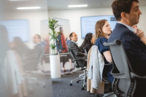 Several adults sit in office chairs around a conference table, attentively listening. The setting is a modern, bright meeting room with a large potted plant, blurred screens, and posters in the background. The scene appears professional and focused.