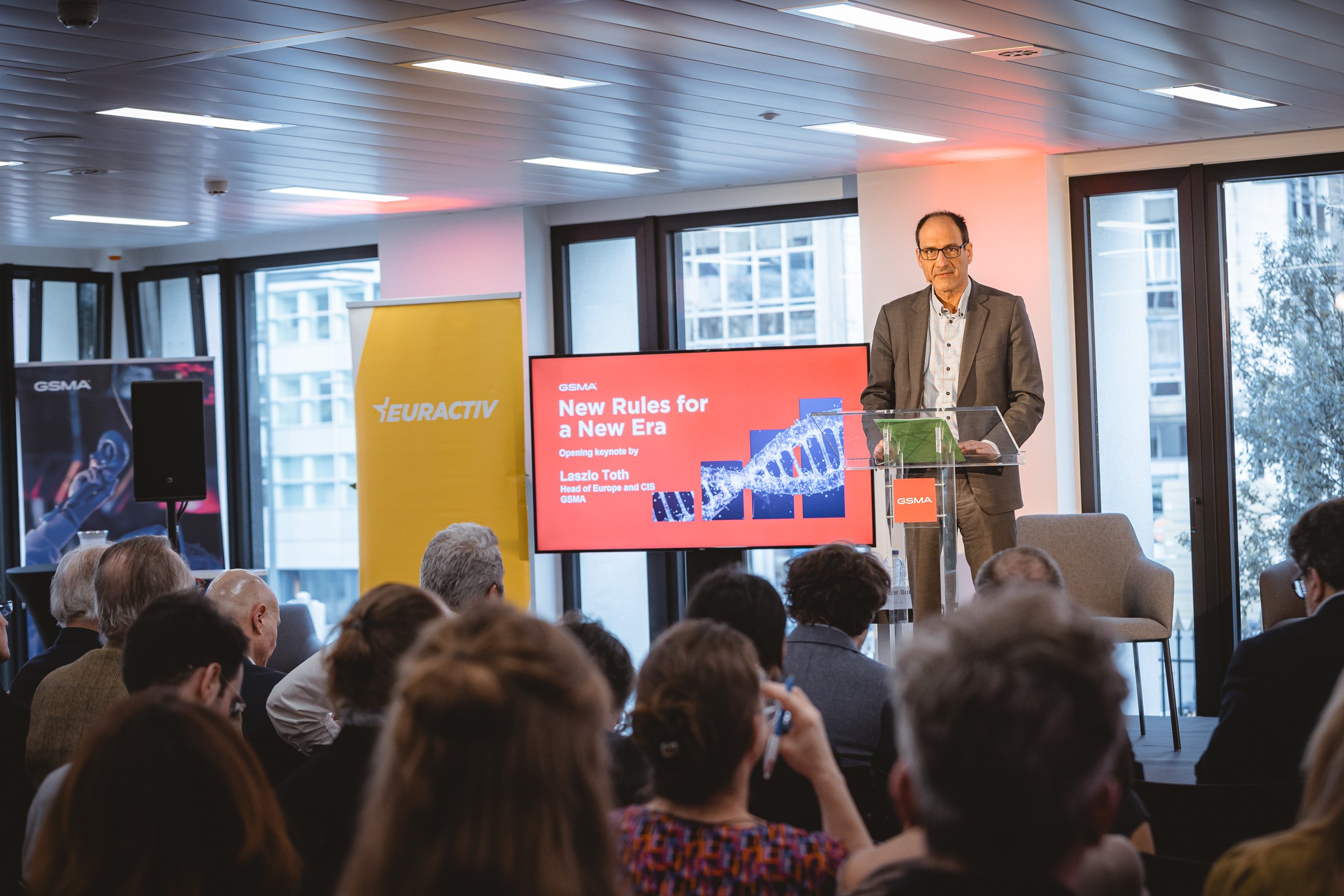 A man in a suit speaks at a podium in front of an audience. Behind him is a screen displaying “New Rules for a New Era” with images of DNA and digital graphics, as well as logos for GSMA and EURACTIV. Large windows and office lights fill the background.
