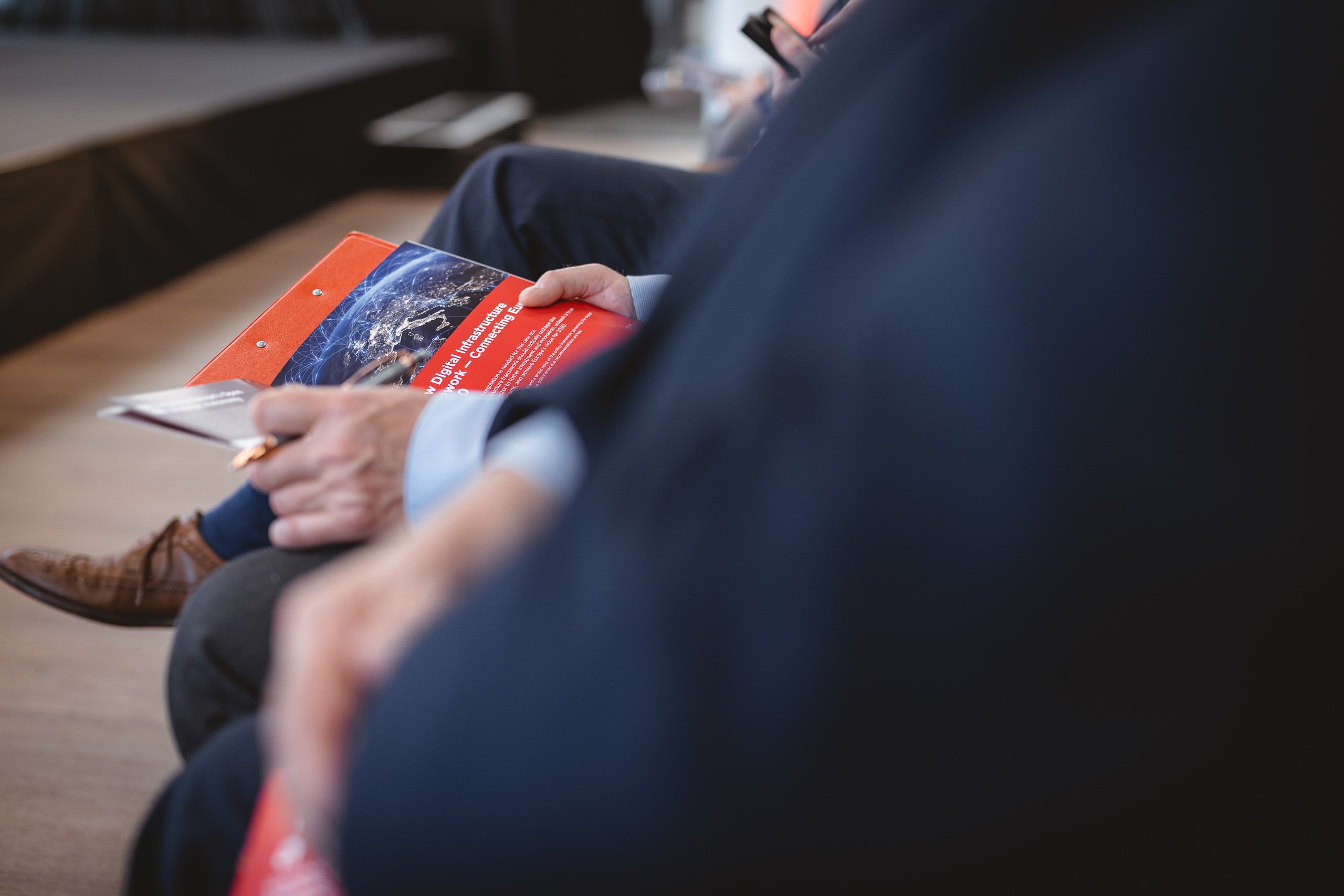 A close-up of a seated person in a suit holding a red booklet, with a pen in hand. The booklet has an image of Earth on the cover. Another person in formal attire is blurred in the foreground, and a stage is visible in the background.