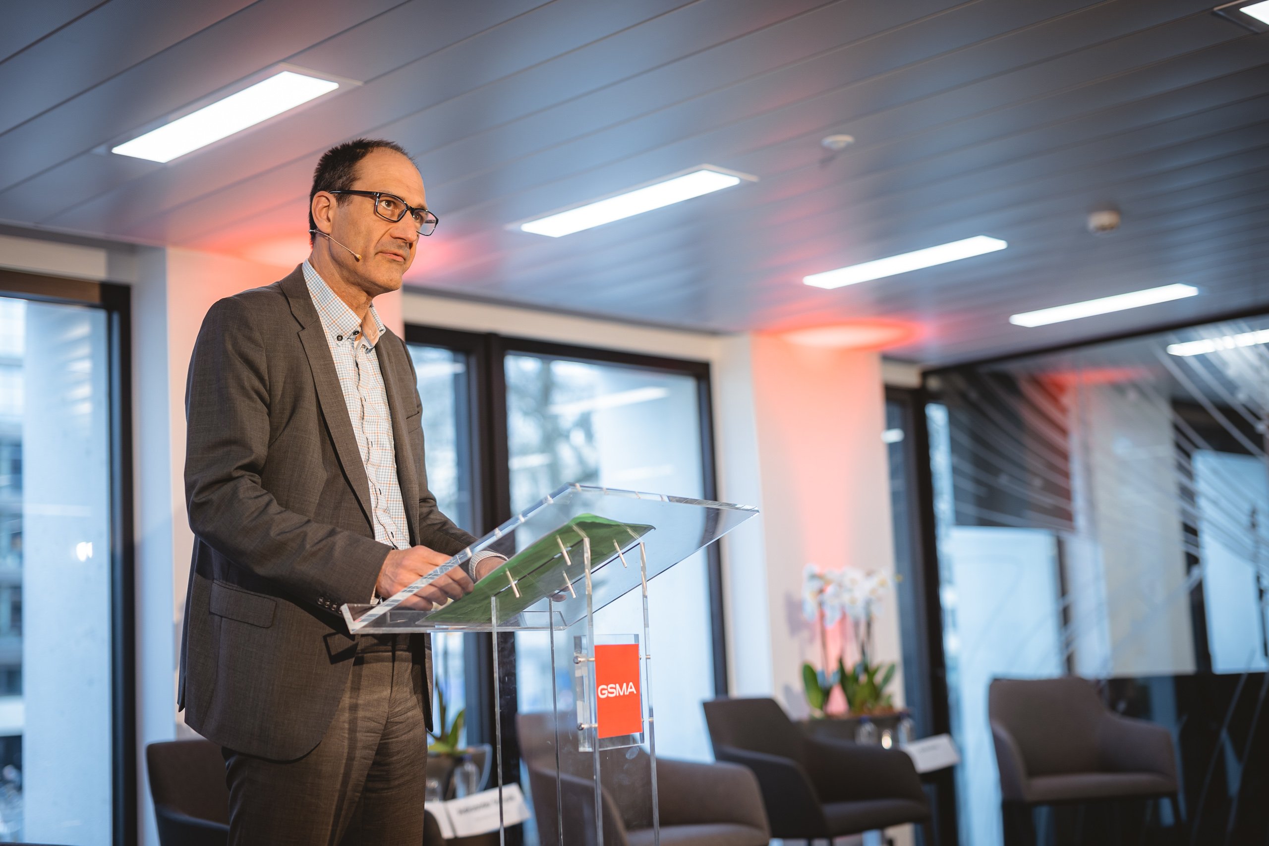 A man in a suit and glasses stands at a clear podium labeled "GSMA," speaking in a modern conference room with large windows, empty chairs, and potted plants in the background. Warm lighting illuminates the scene.