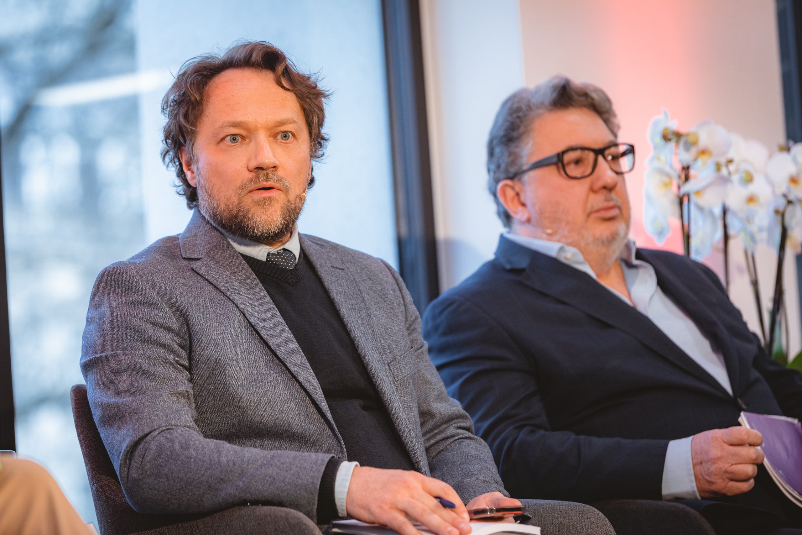 Two men in suits sit next to each other indoors. The man on the left has wavy brown hair and a beard, wearing a grey suit and black sweater. The man on the right has gray hair, glasses, and a dark suit. A window and white orchids are in the background.