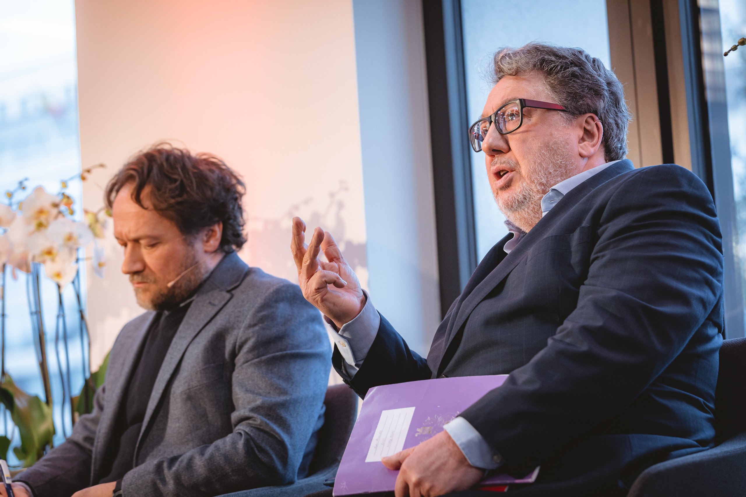 Two men in suits sit side by side in a modern indoor setting. The man on the left looks down, listening intently, while the man on the right speaks and gestures with his hand, holding a purple folder. Soft lighting and potted orchids appear in the background.