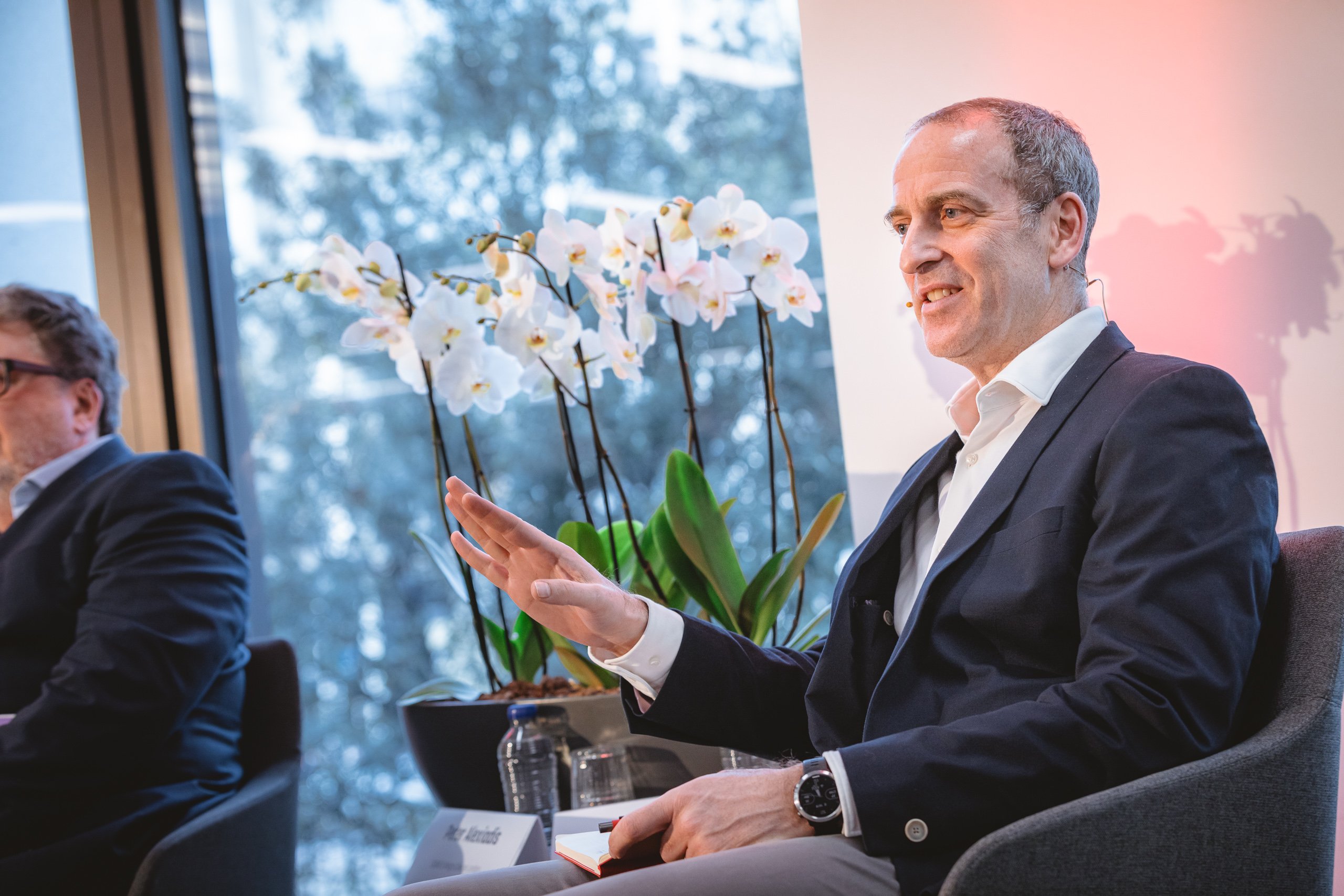 A man in a suit sits in a chair, gesturing with his hand while talking. He smiles and wears a smartwatch. White orchids and large windows with trees outside are in the background. Another man is partly visible, seated to the left.