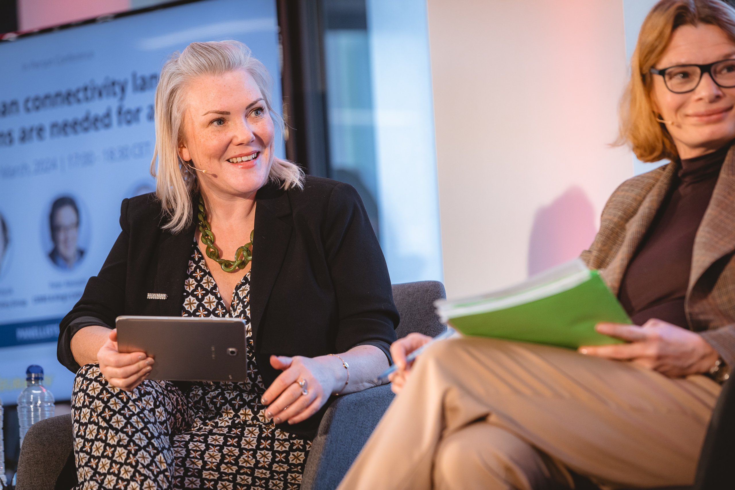 Two women are seated and engaged in conversation at an event. The woman on the left has light hair, wears a black blazer and patterned dress, and holds a tablet. The woman on the right has glasses, a brown blazer, and holds green folders. A blurred presentation screen is behind them.