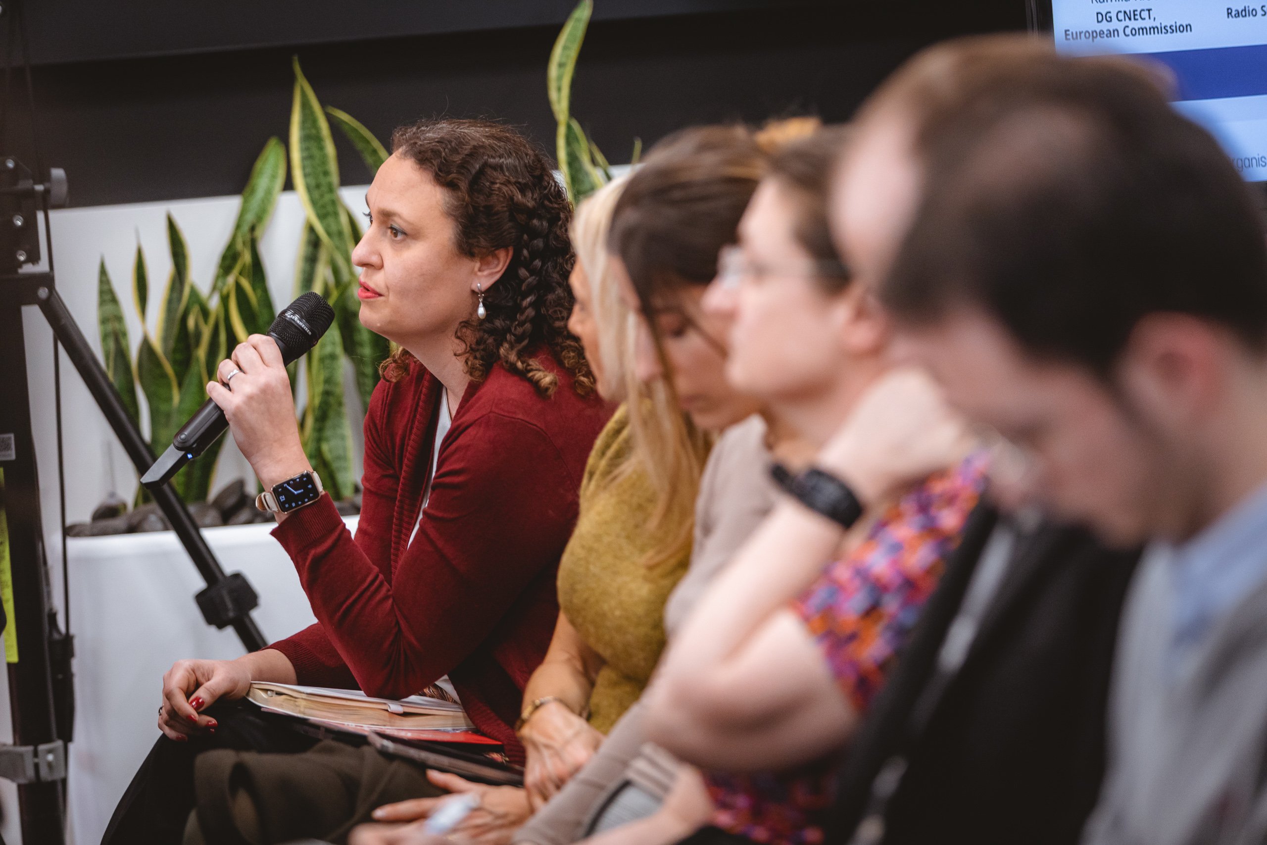 A woman with curly brown hair and a red cardigan speaks into a microphone while seated in a row with four other people, who appear to be listening attentively. Green potted plants and a partially visible presentation screen are in the background.