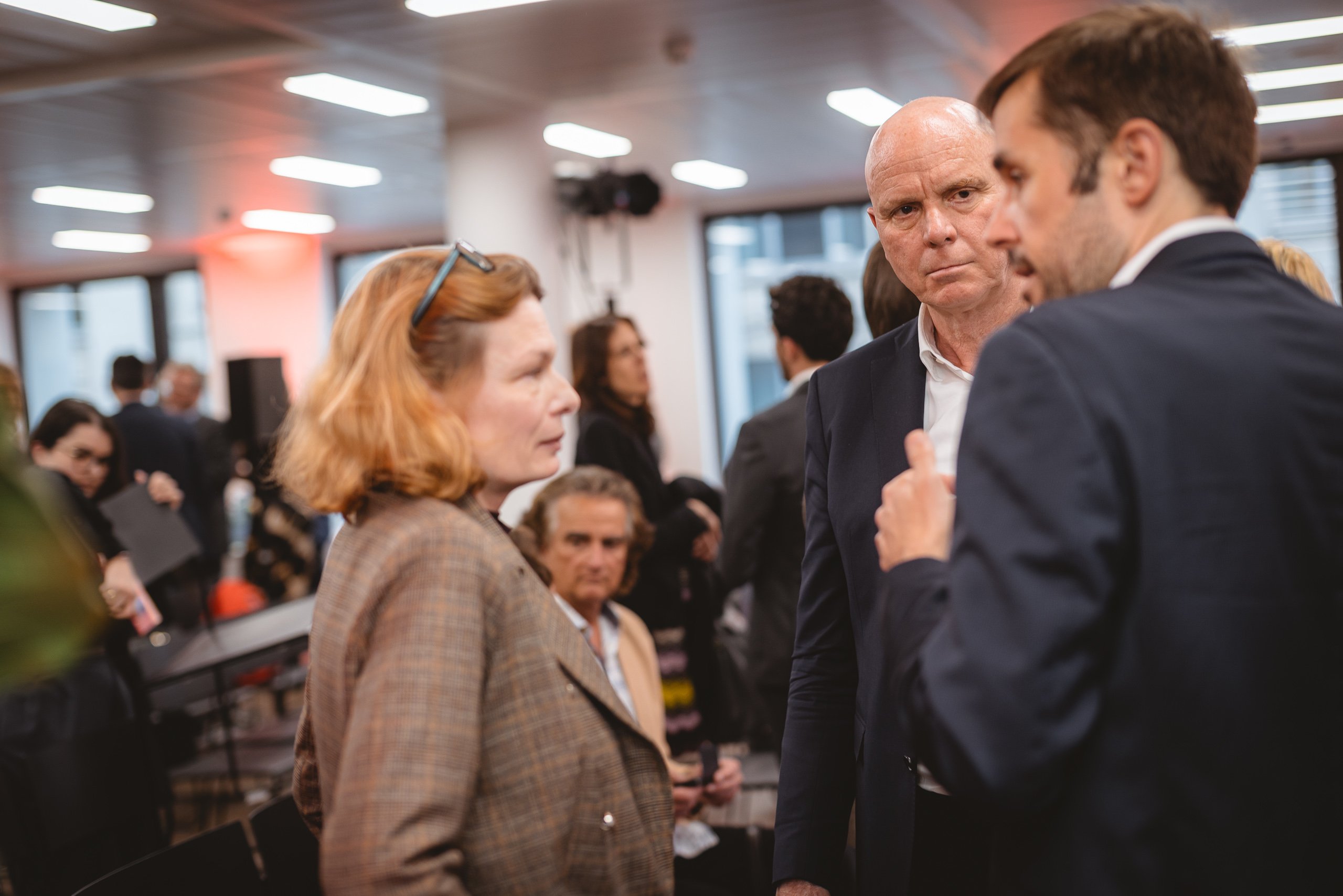 A woman in a brown blazer listens to two men in suits talking at an indoor event. The man in the center looks serious, while others converse in the blurred background under bright overhead lights. The scene appears professional or corporate.