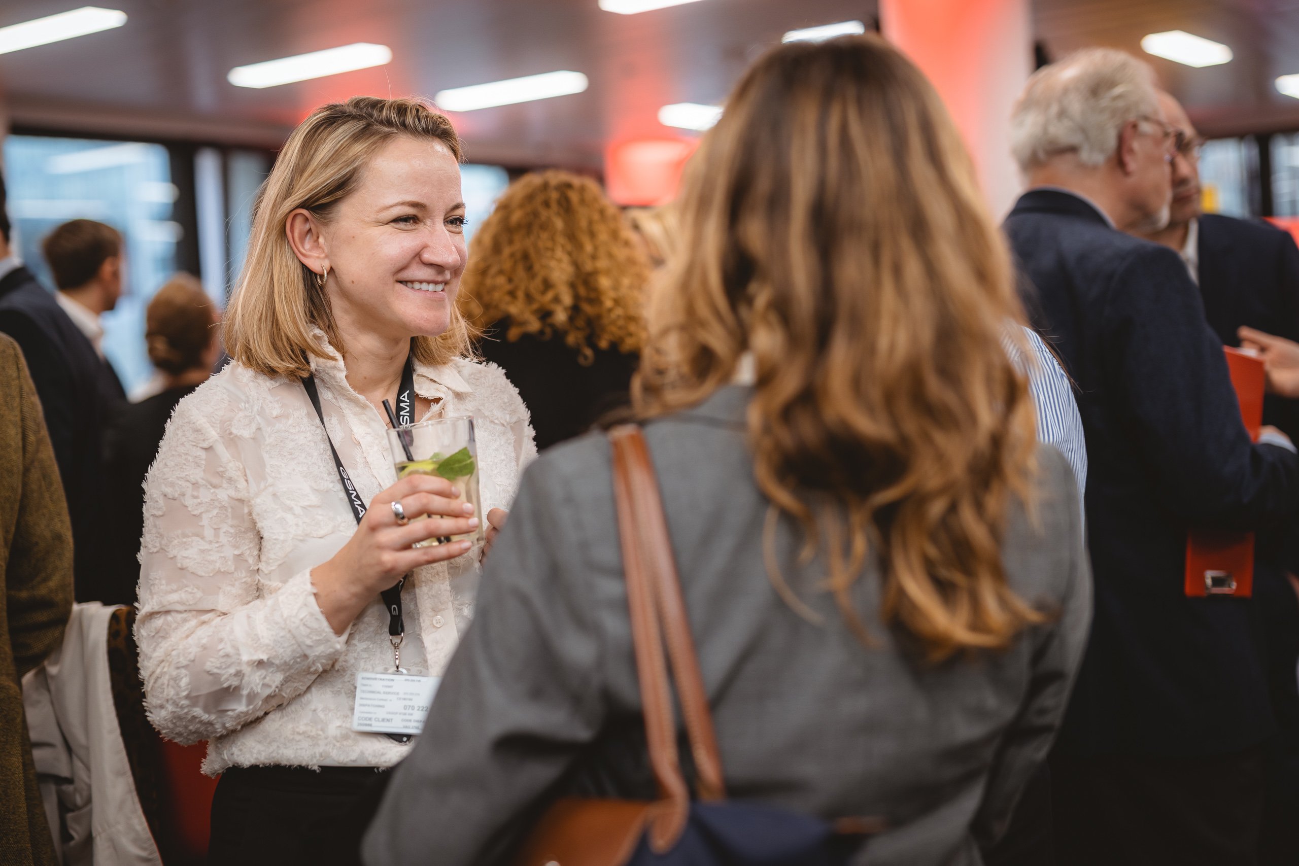 A woman in a white blouse smiles and holds a drink while talking to another woman with light brown hair at a busy indoor networking event. People in business attire are gathered in the background under warm lighting.