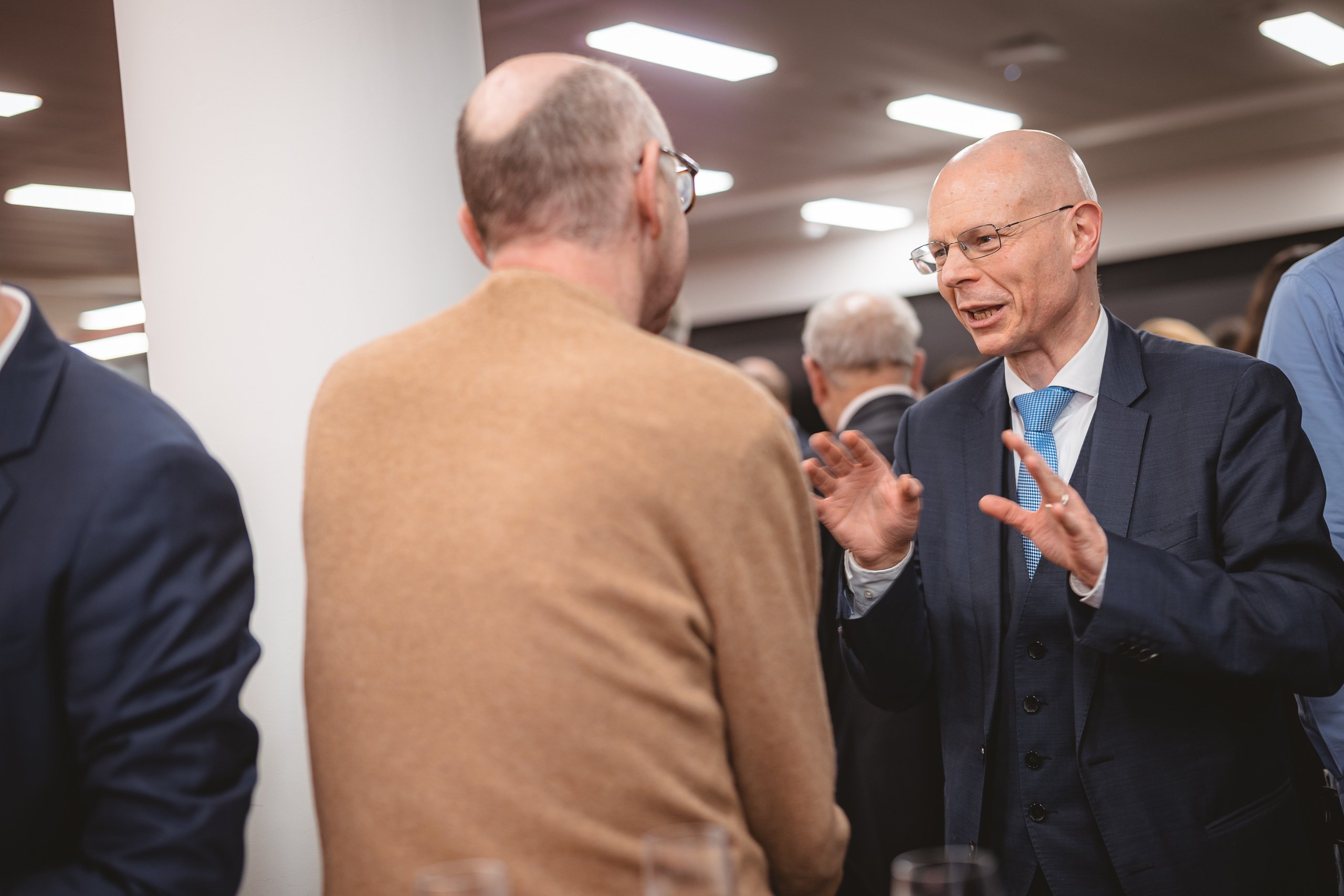 Two men, both bald and wearing glasses, are engaged in conversation at an indoor event. One is in a suit and tie, gesturing animatedly, while the other, in a tan sweater, listens. Other people and bright overhead lights are visible in the background.