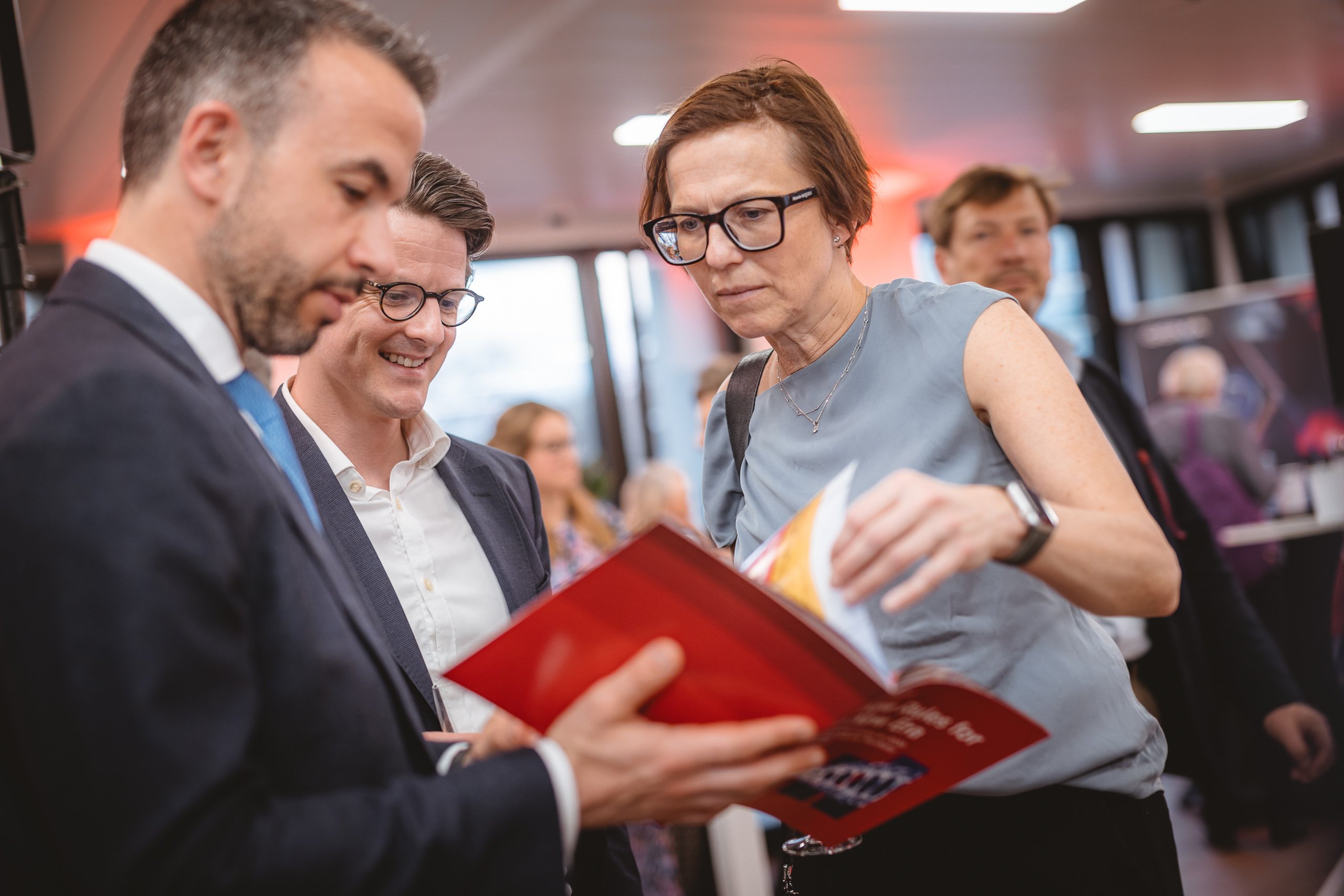 Three people stand indoors at an event. A man in a suit holds and shows a red booklet as another man in glasses and a woman with short hair and glasses look on intently. Other people and display boards are visible in the background.