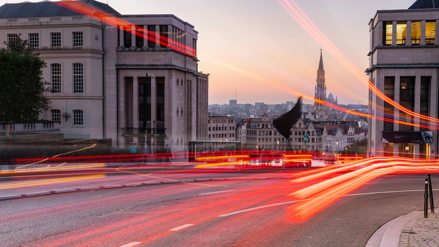 A city street at dusk with light trails from moving cars, flanked by two large stone buildings. In the background, a tall, ornate spire rises above the city skyline under a pastel-colored sky. The scene feels dynamic and urban.